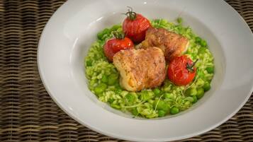 A white plate with green peas, cherry tomatoes, and a piece of cooked meat on a woven mat.