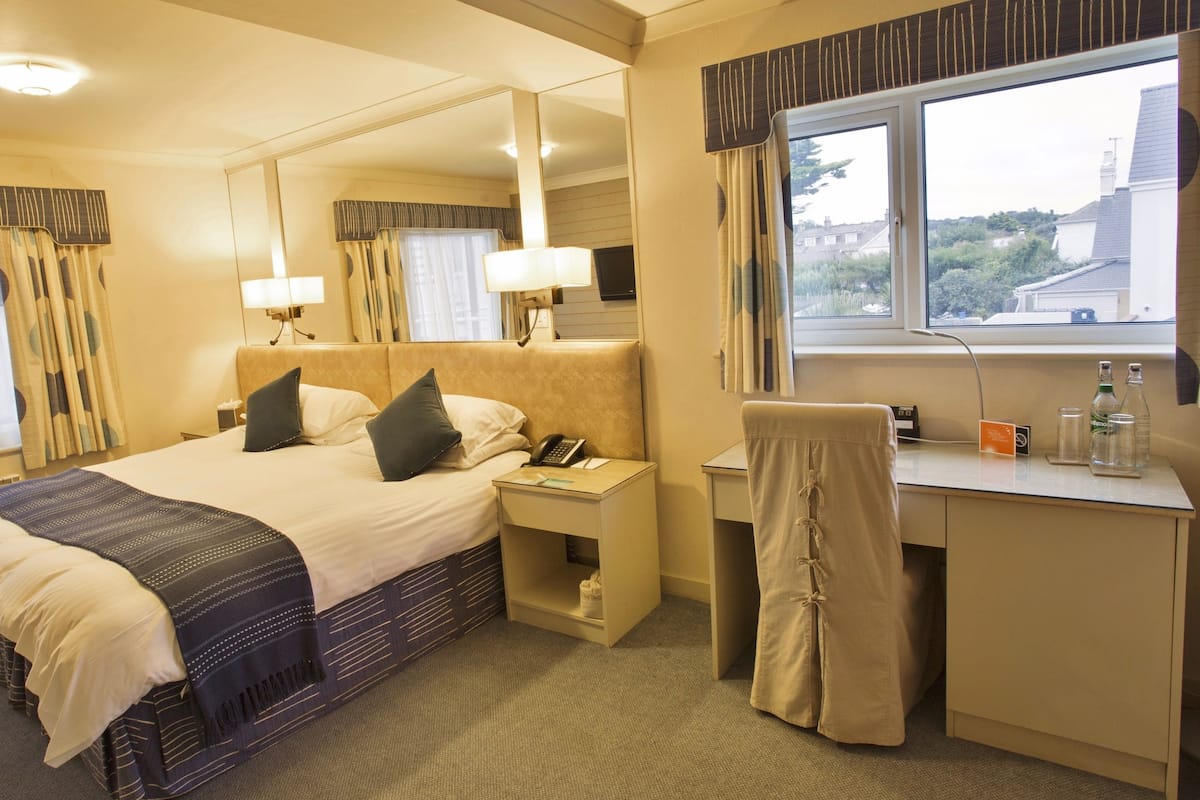 Une chambre d’hôtel équipée d’un lit, d’une table de chevet, d’un bureau, d’une chaise et d’une fenêtre donnant sur des immeubles.