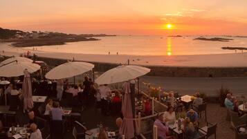 An outdoor dining area with tables and umbrellas overlooking a beach at sunset.