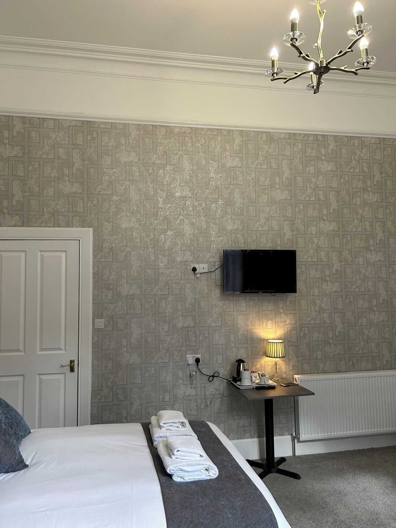 A hotel room with a bed, a TV mounted on the wall, a bedside table with a lamp, and a radiator.
