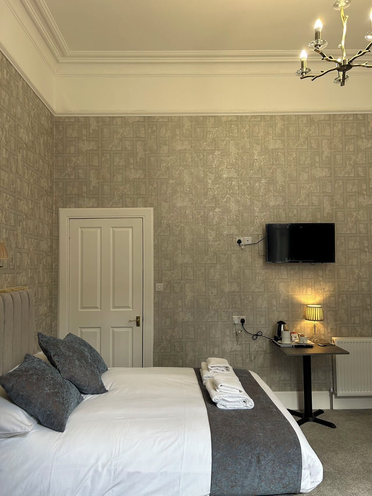 A hotel room with a bed, a TV mounted on the wall, a bedside table with a lamp, and a door.
