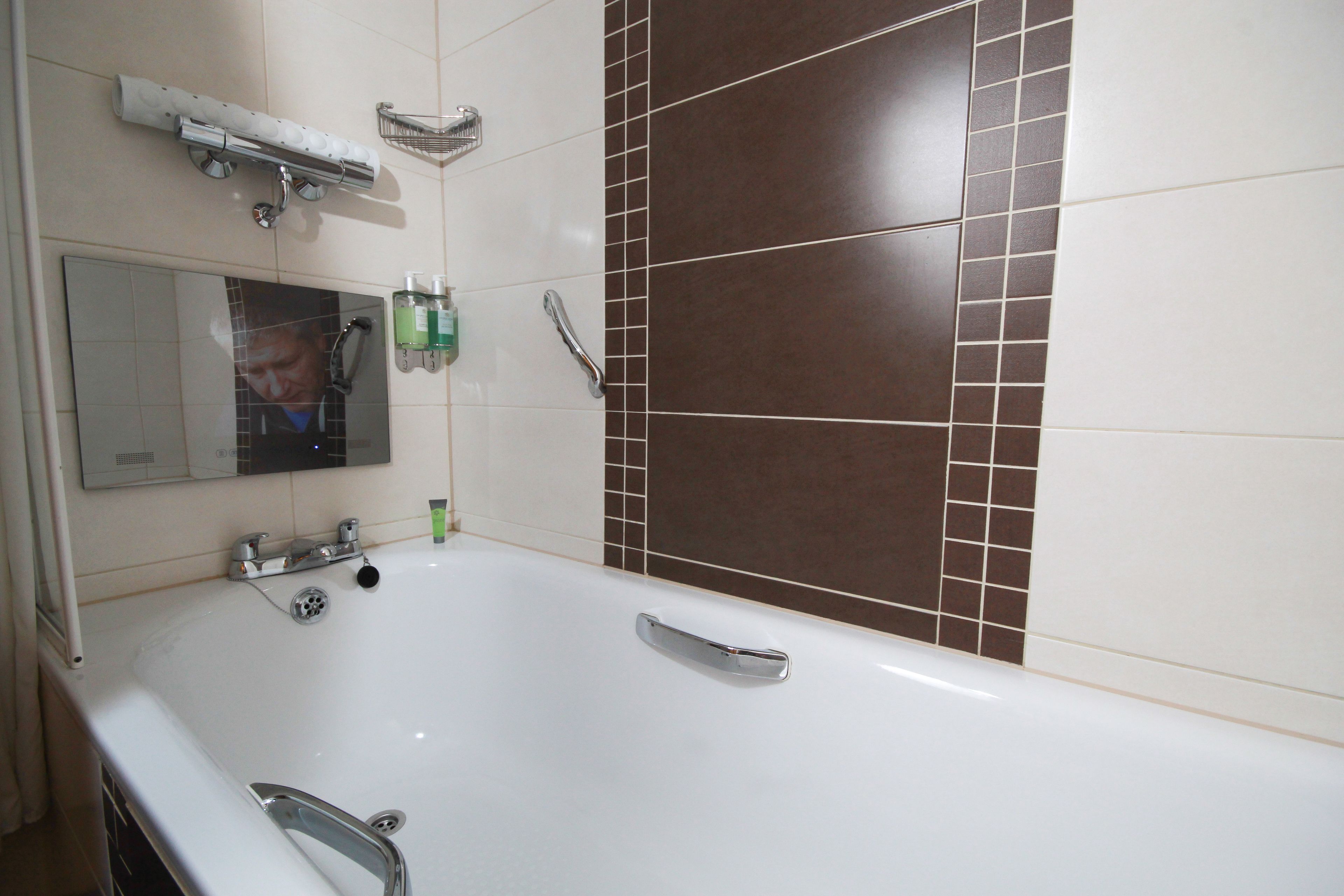 A bathroom with a large white bathtub, a mirror, and brown tiled walls.