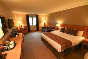 Habitación de hotel con una cama grande, dos mesitas de noche, un escritorio con silla, un televisor y una ventana con cortinas.