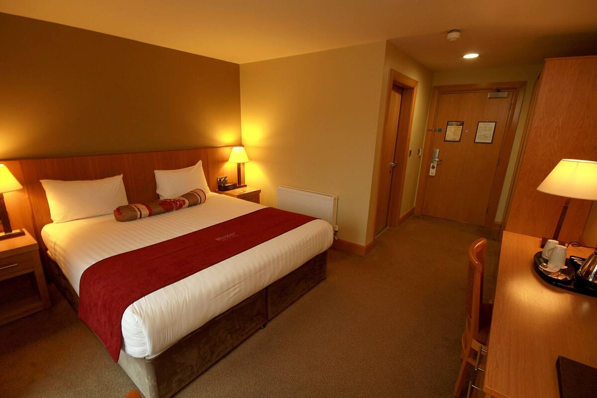 Ein Hotelzimmer mit einem großen Bett, zwei Nachttischen mit Lampen, einem Holz-Kopfteil, einem Holzschrank und einer Tür zu einem weiteren Raum.