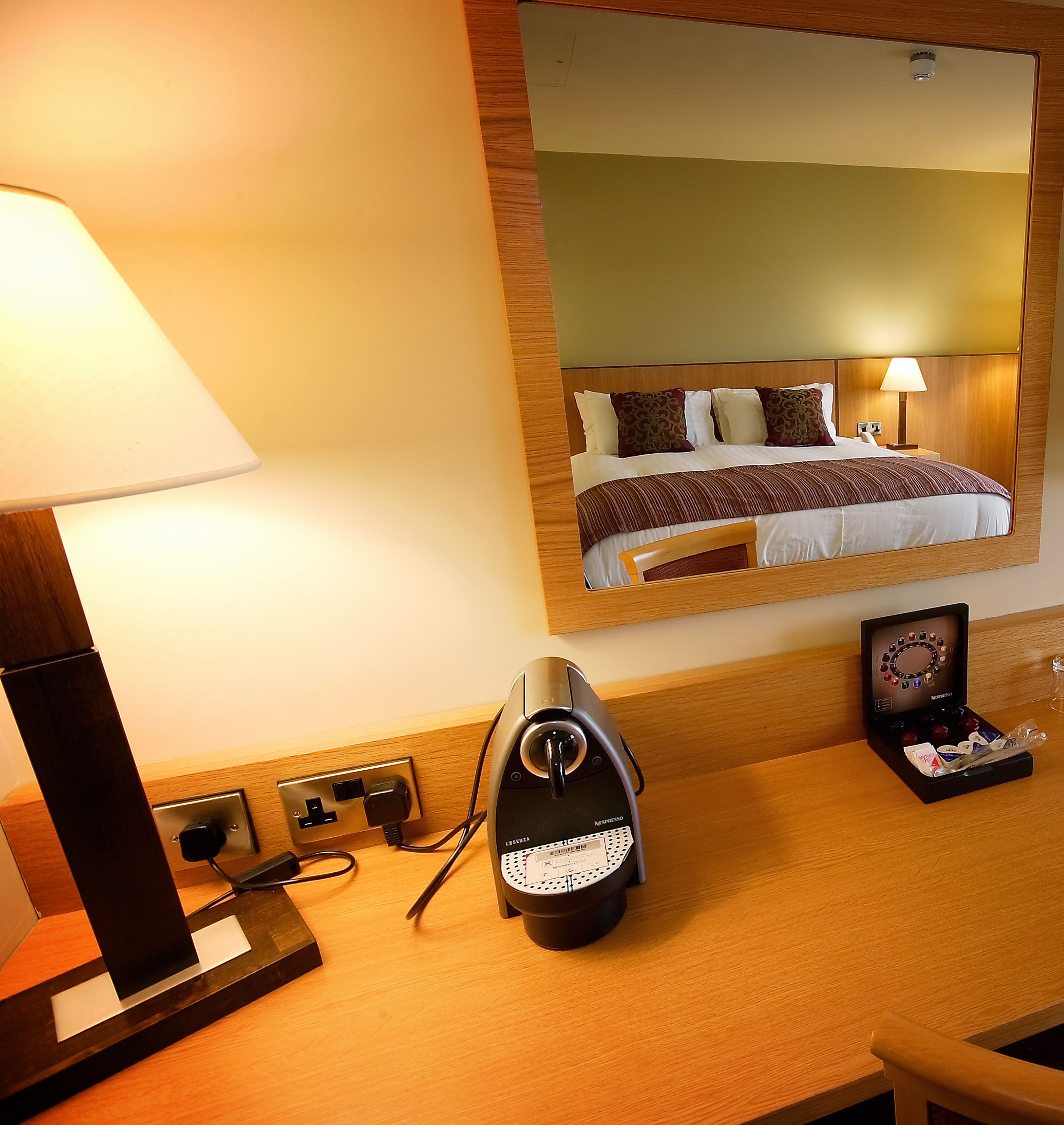 A hotel room with a bed, a bedside lamp, a coffee maker, and a mirror reflecting the room.
