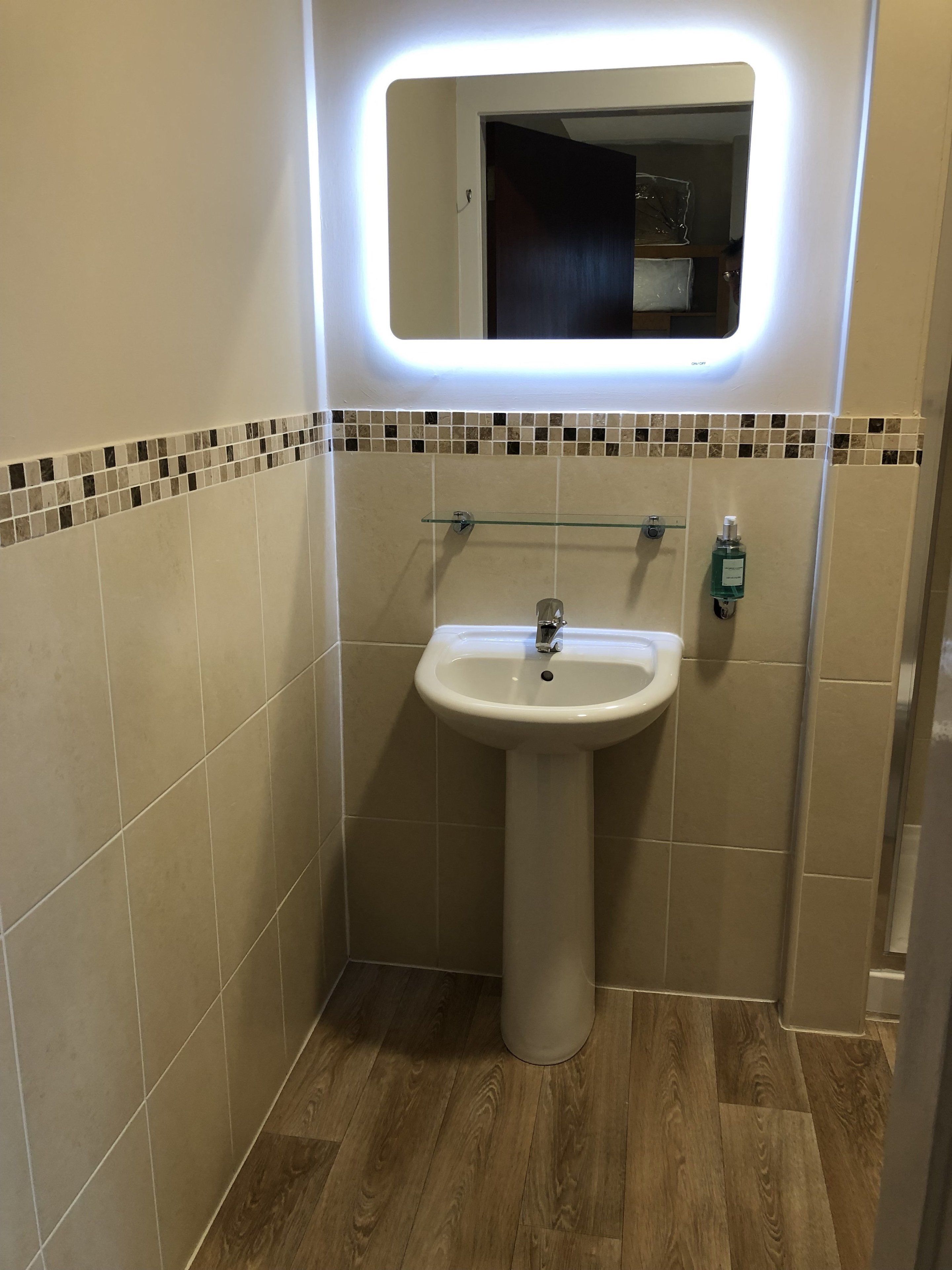 A bathroom with a pedestal sink, a mirror with a light, and a tiled backsplash.