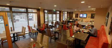 A restaurant with multiple tables and booths, large windows, and a television screen on the wall.