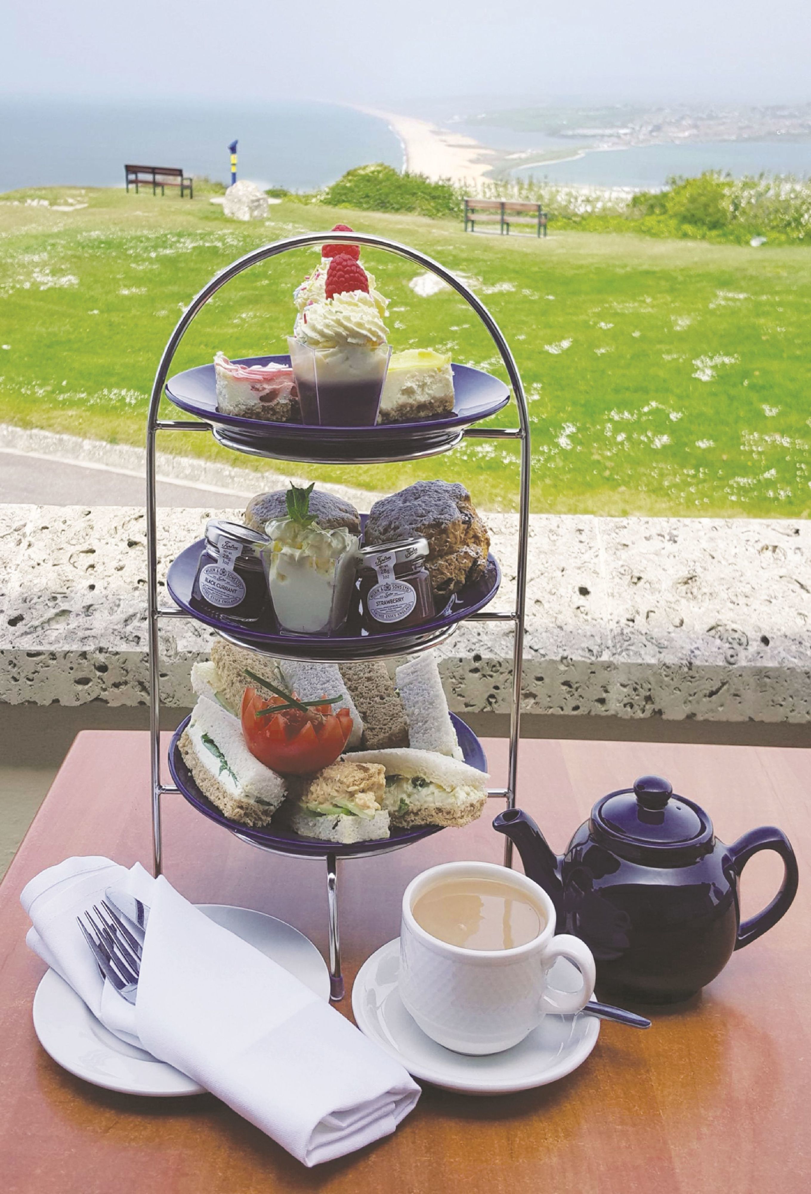 Un expositor escalonado para postres con sándwiches y pasteles, una tetera y una taza de té sobre una mesa con vistas panorámicas.