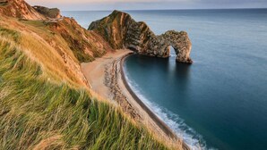 Un paysage côtier caractérisé par une arche rocheuse naturelle, une plage de sable et des falaises herbeuses.