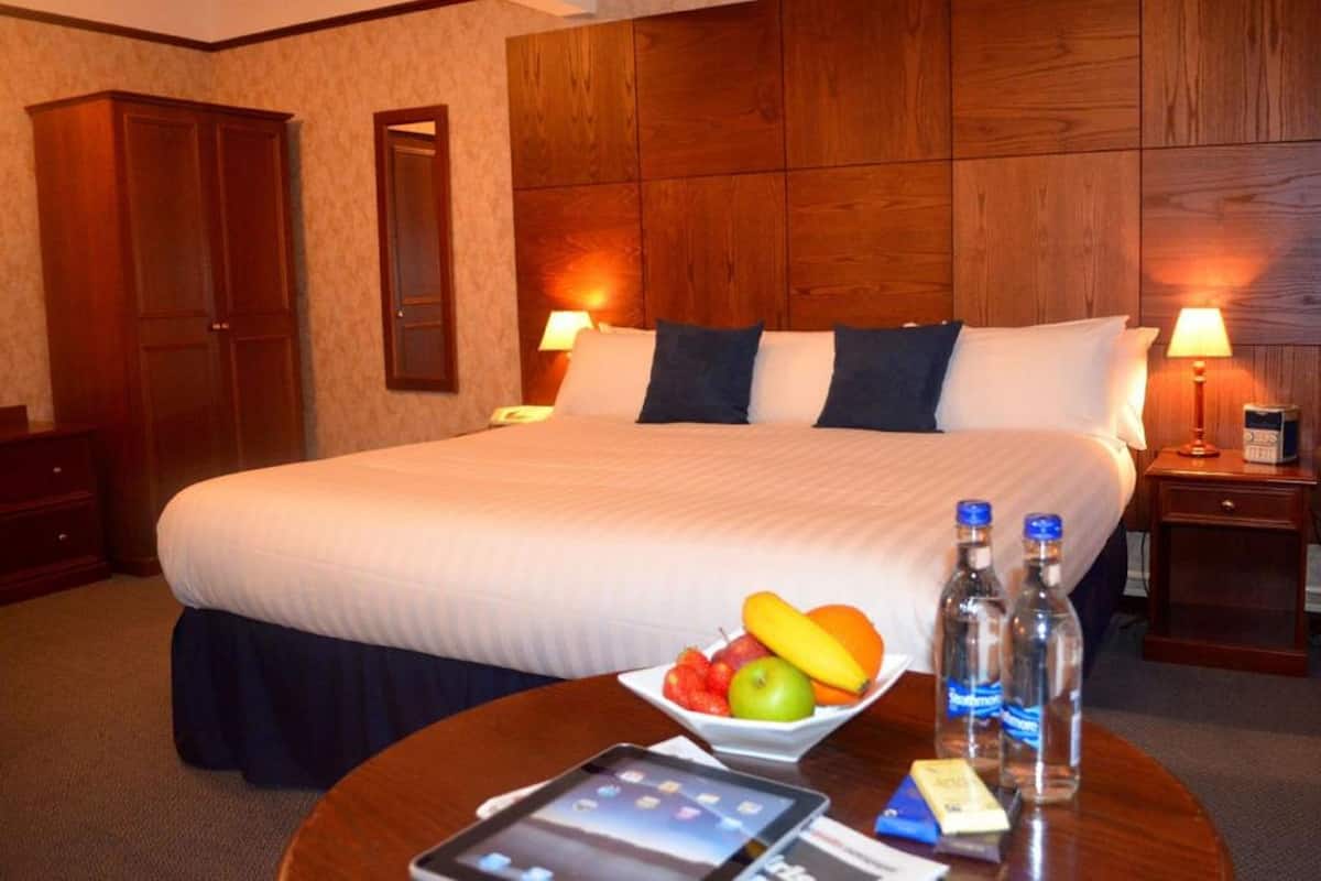 A hotel room with a large bed, wooden headboard, two bedside tables with lamps, a bedside table with fruit and a bottle, and a tablet on the floor.
