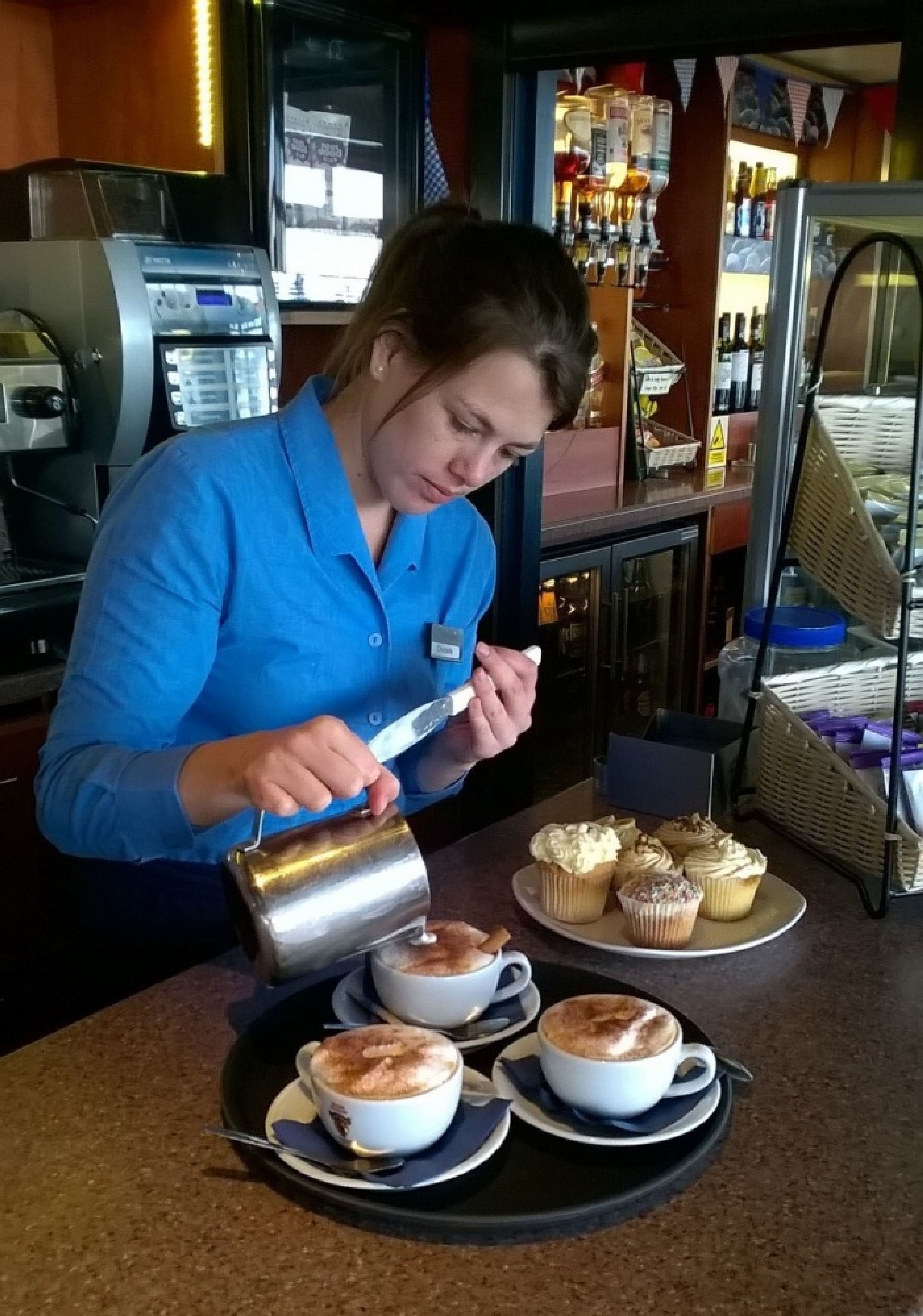 A person in a blue shirt pouring a beverage into a cup. There are several cups and a tray with cupcakes on a counter.