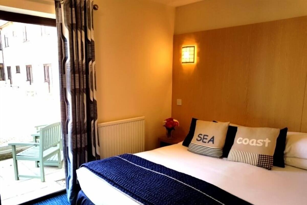 A hotel room with a bed, a window with curtains, a small table with a vase, and a view of the outside balcony.