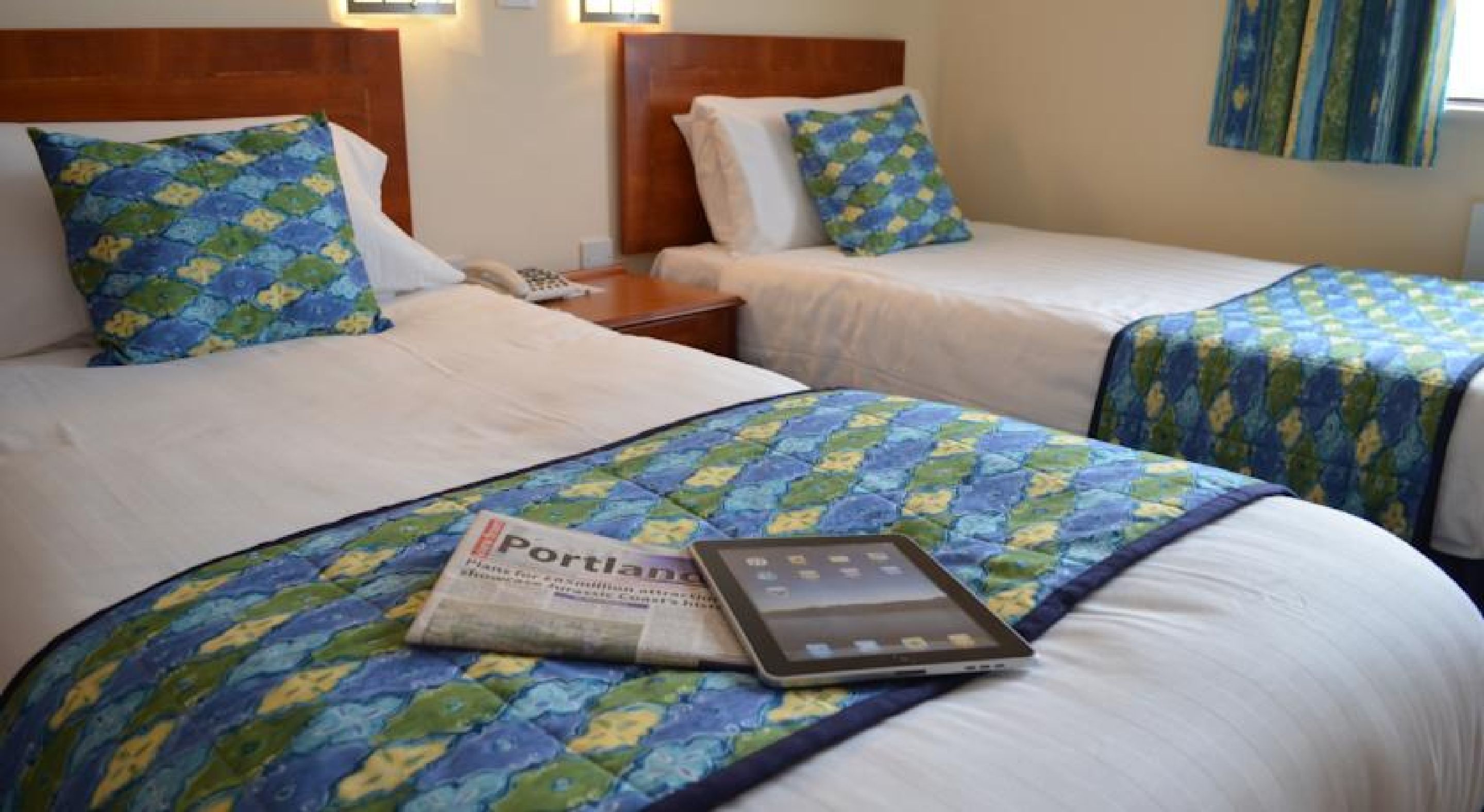 A hotel room with two beds, each with a blue and green patterned bedspread, a bedside table with a lamp, and a tablet on the bed.
