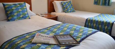 A hotel room with two beds, each with a blue and green patterned bedspread, a bedside table with a lamp, and a tablet on the bed.