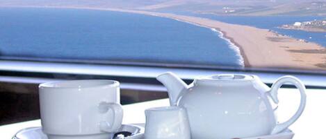 A table with a teapot, cups, and cookies, overlooking a beach and ocean.