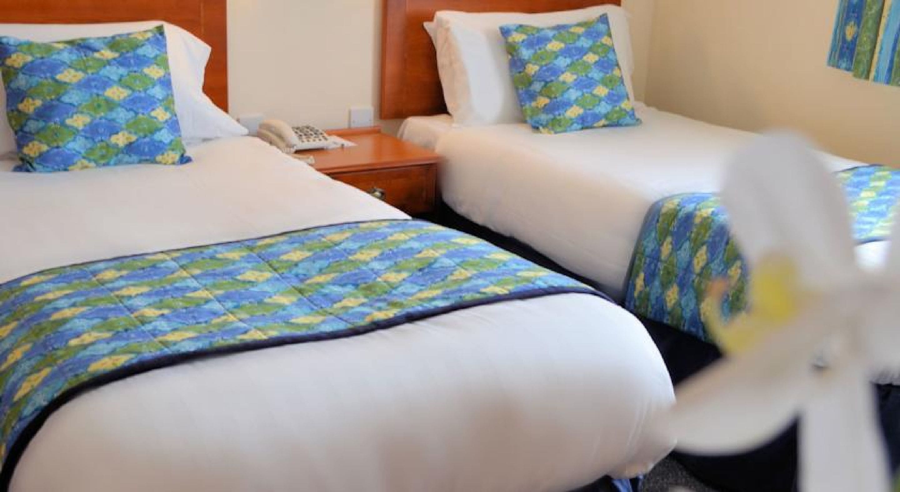 Une chambre d’hôtel avec deux lits, chacun recouvert d’une couvre-lit à motifs bleus et verts, assortie à des coussins du même style. Une table de chevet en bois avec un téléphone se trouve entre les deux lits.
