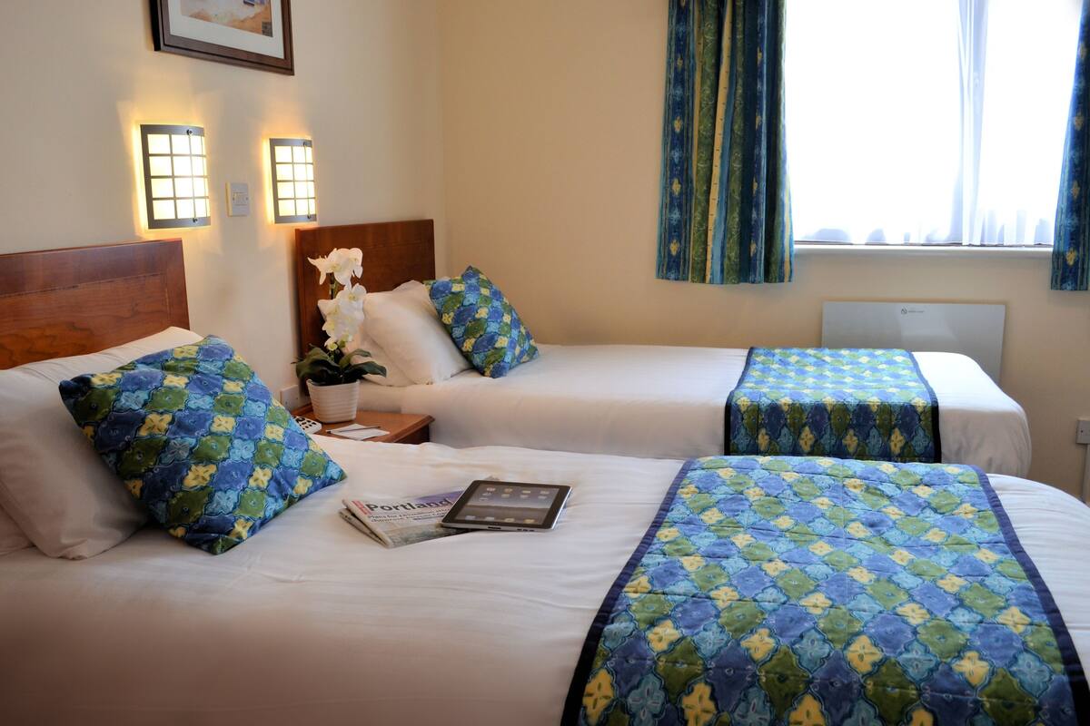 A hotel room with two beds, each with a blue and green patterned bedspread, white pillows, and a wooden headboard. There is a nightstand with a lamp, a potted plant, and a tablet on one bed. The room has a window with curtains and a framed picture on the wall.