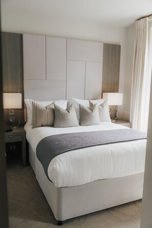 A hotel room with a large bed, two bedside tables with lamps, a headboard, and a window with curtains. - Dunalastair Hotel Suites (Pitlochry)
