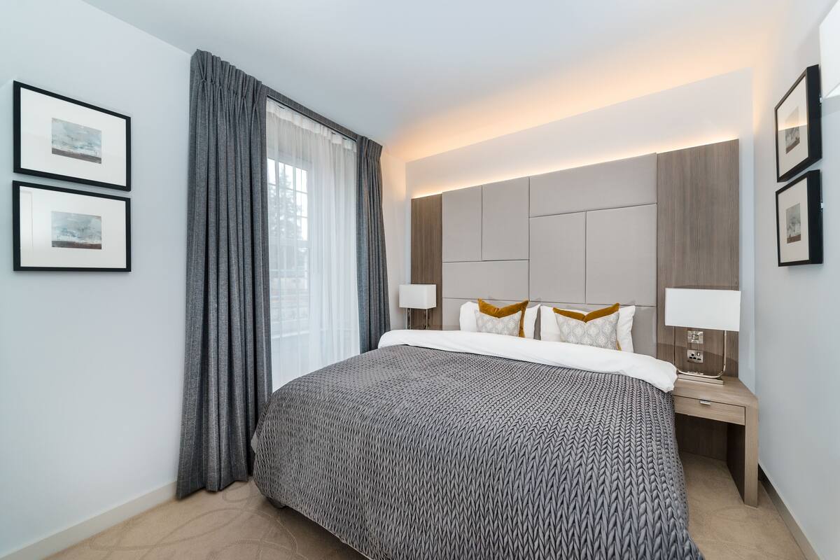 Quarto de hotel moderno com uma cama grande, uma mesa de cabeceira com abajur e duas imagens emolduradas na parede.