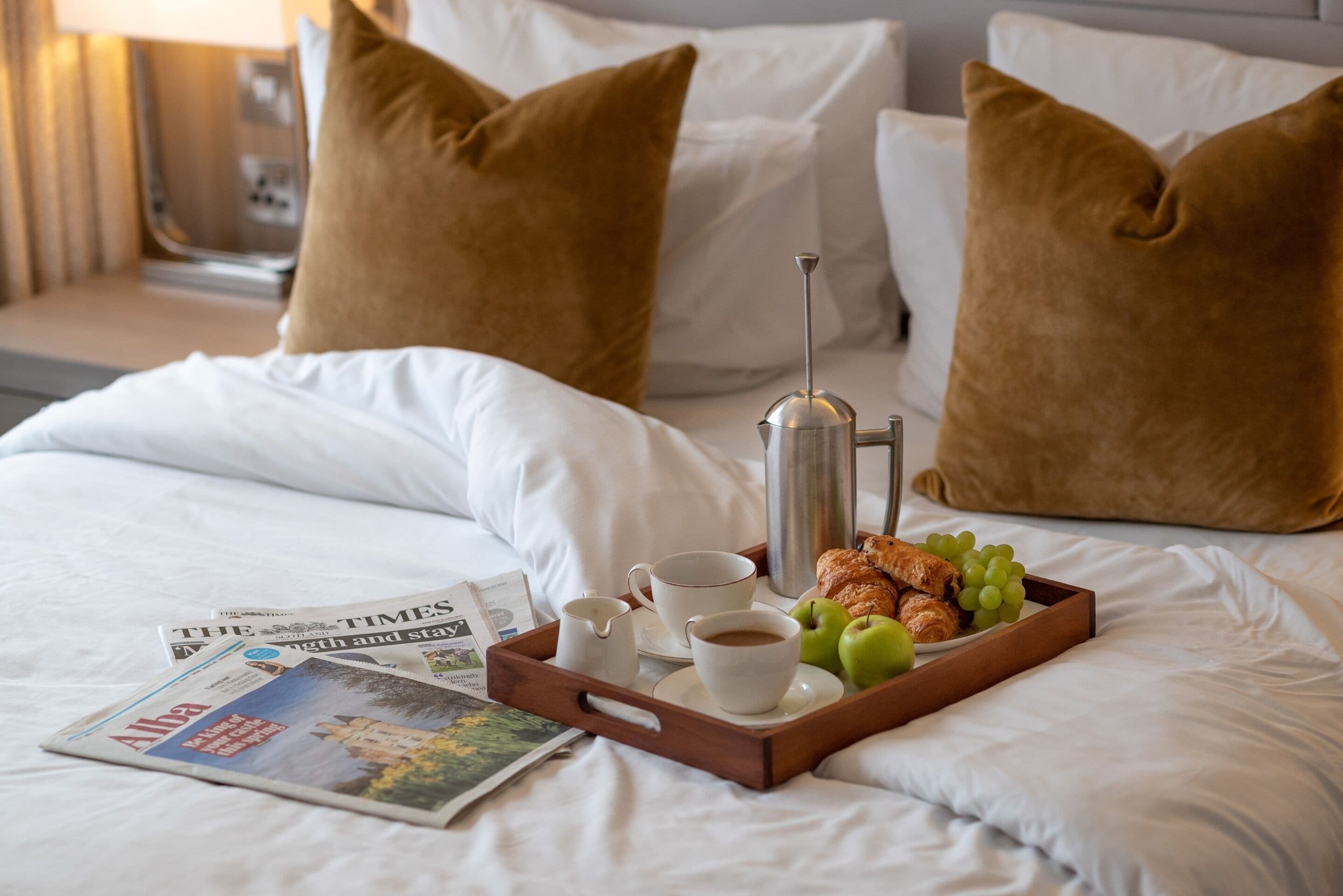 A neatly made bed with white linens, a tray with breakfast items including croissants, apples, and grapes, and a newspaper on the bed.