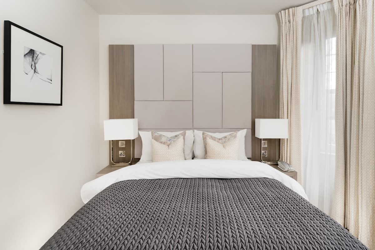Quarto de hotel moderno com uma cama grande, duas mesas de cabeceira com abajures, um quadro na parede e uma janela com cortinas.