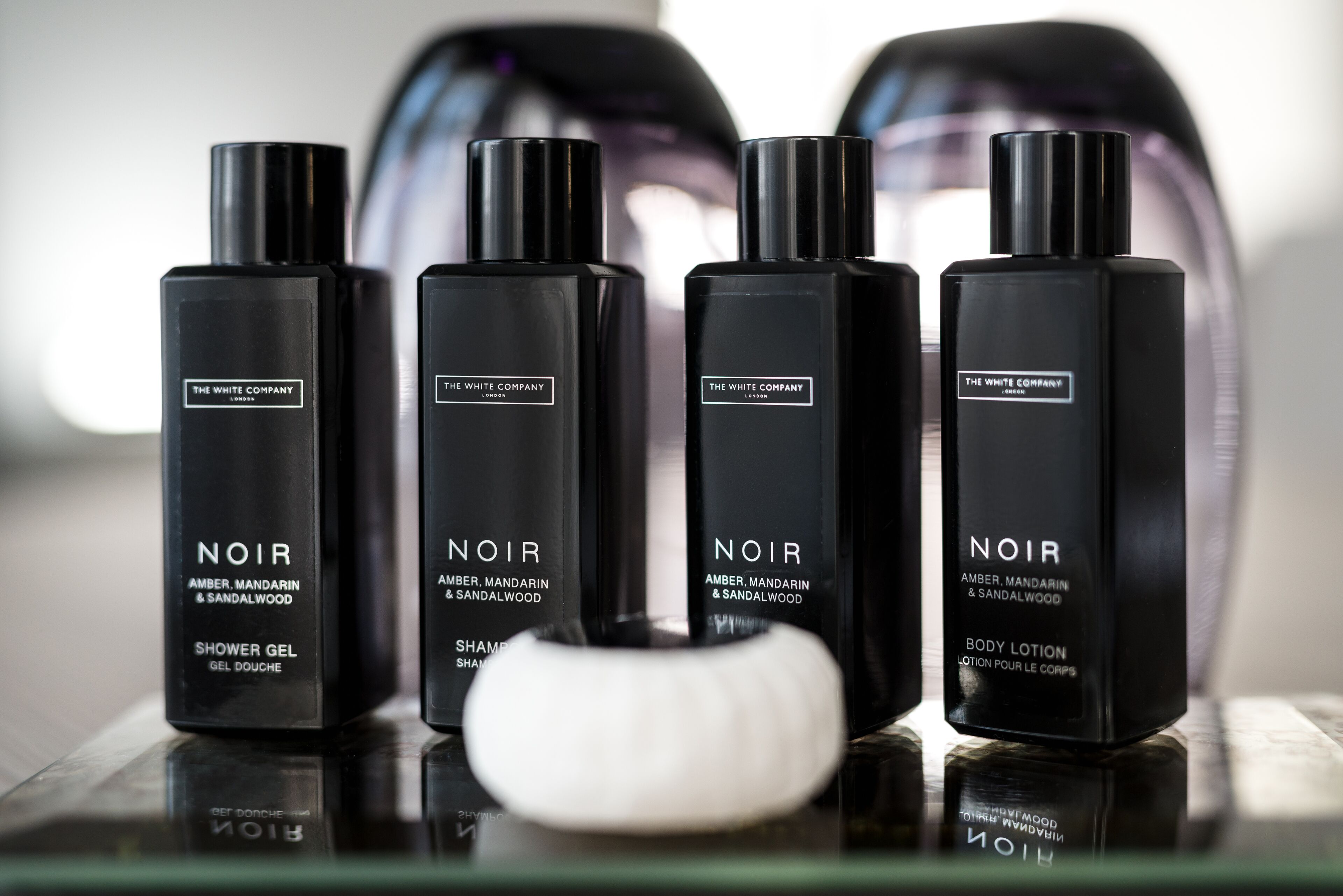 Fire sorte flasker med produkter fra The White Company Noir på en spejlenende overflade.