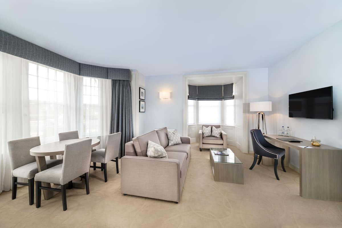 Quarto de hotel moderno com área de estar, mesa de jantar, sofá e televisão fixada na parede.