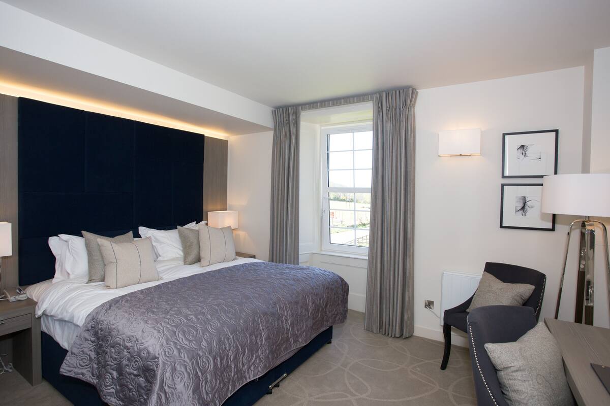 Quarto de hotel com uma cama grande, abajures, uma janela com cortinas e uma cadeira.