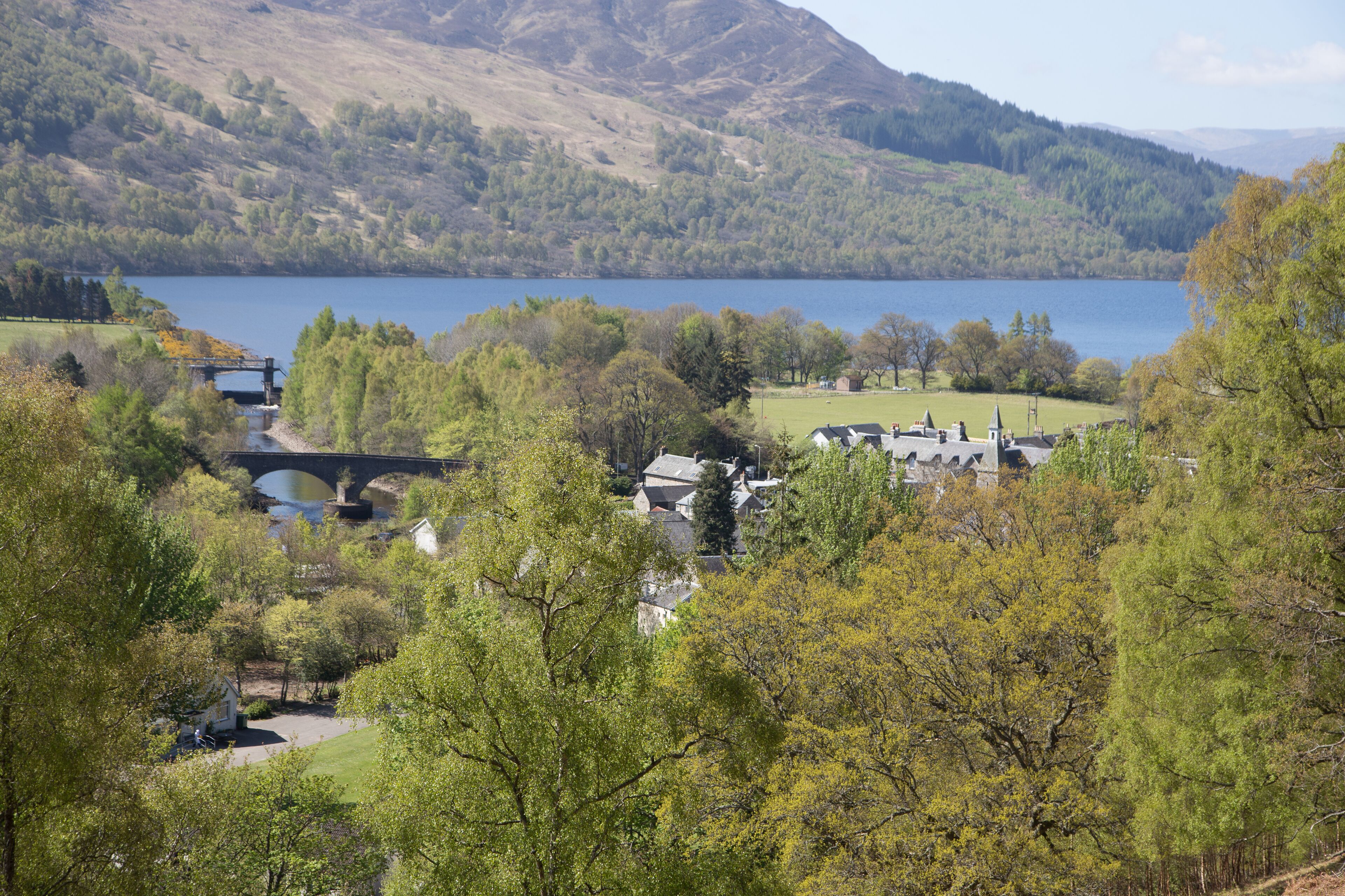 Un village au bord d’un lac, avec un pont, des maisons et des collines environnantes.