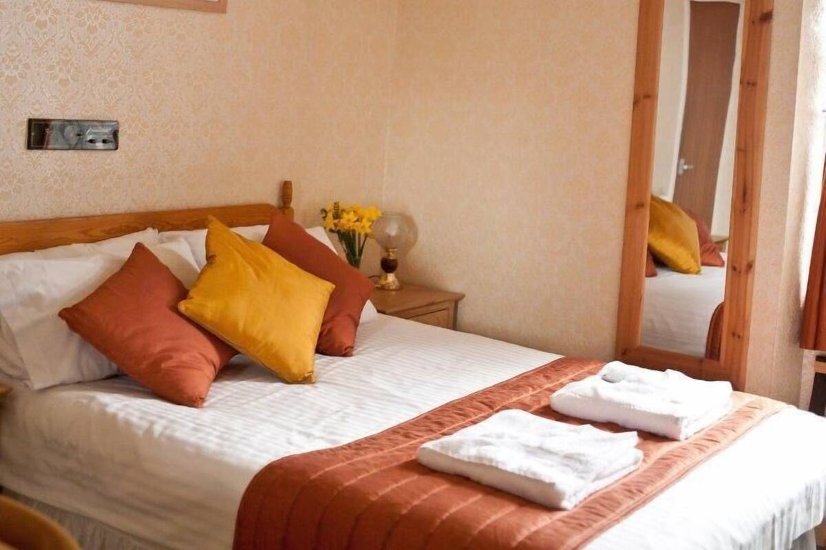 A neatly made bed with white and orange bedding, a bedside table with a vase of yellow flowers, and a mirror on the wall.