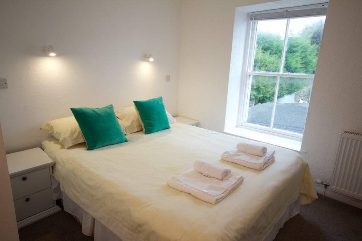 A bedroom with a bed, two pillows, a nightstand, and a window with a view of greenery.