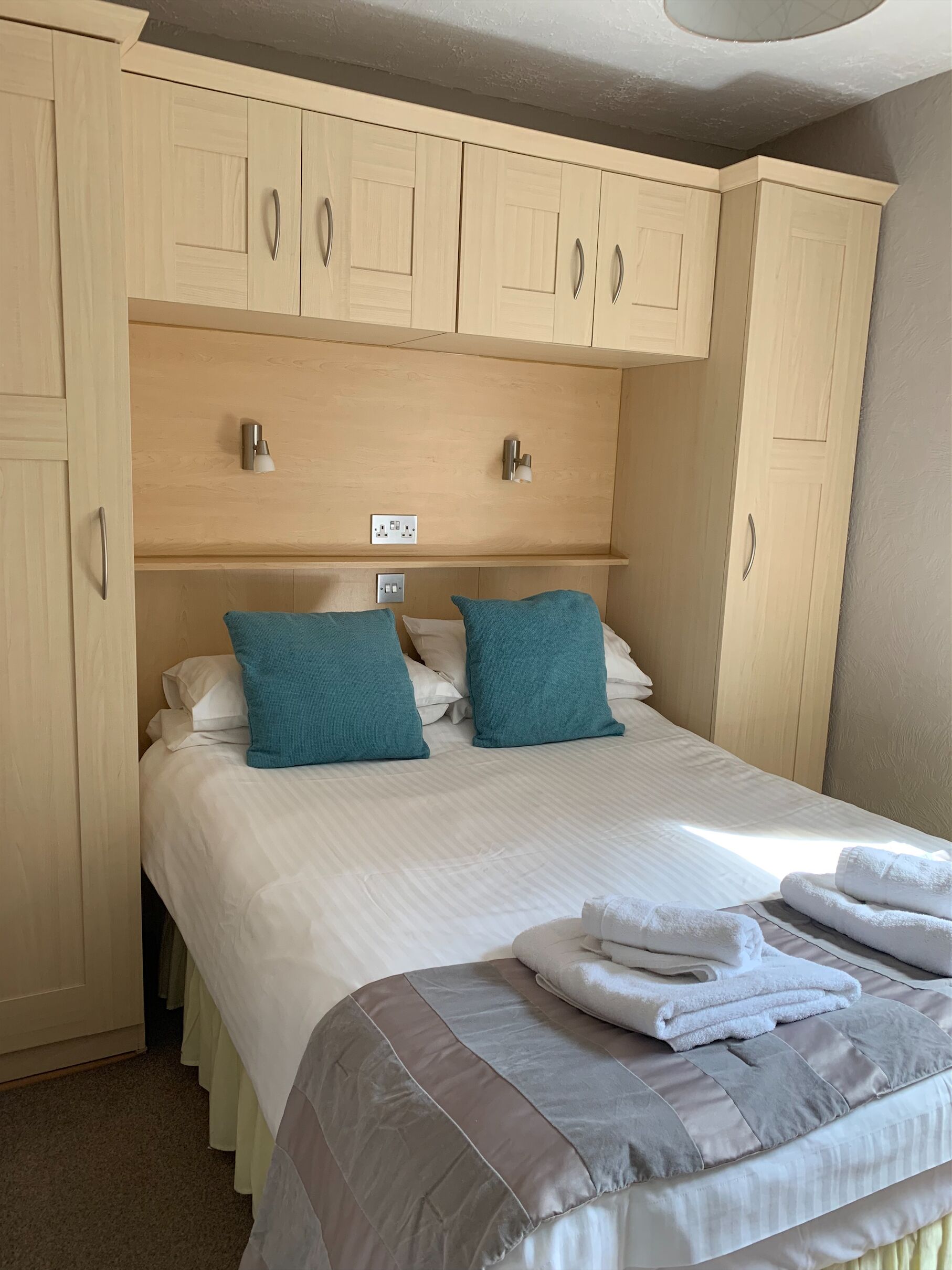 Habitación de hotel con cama, dos almohadas color turquesa, edredón blanco y gris, y un juego de toallas blancas.