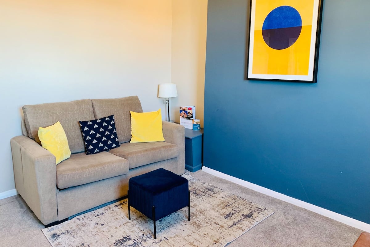 A living room with a beige sofa, two yellow and one dark blue pillow, a small dark blue ottoman, a patterned rug, a lamp, and a framed abstract painting on a teal wall.