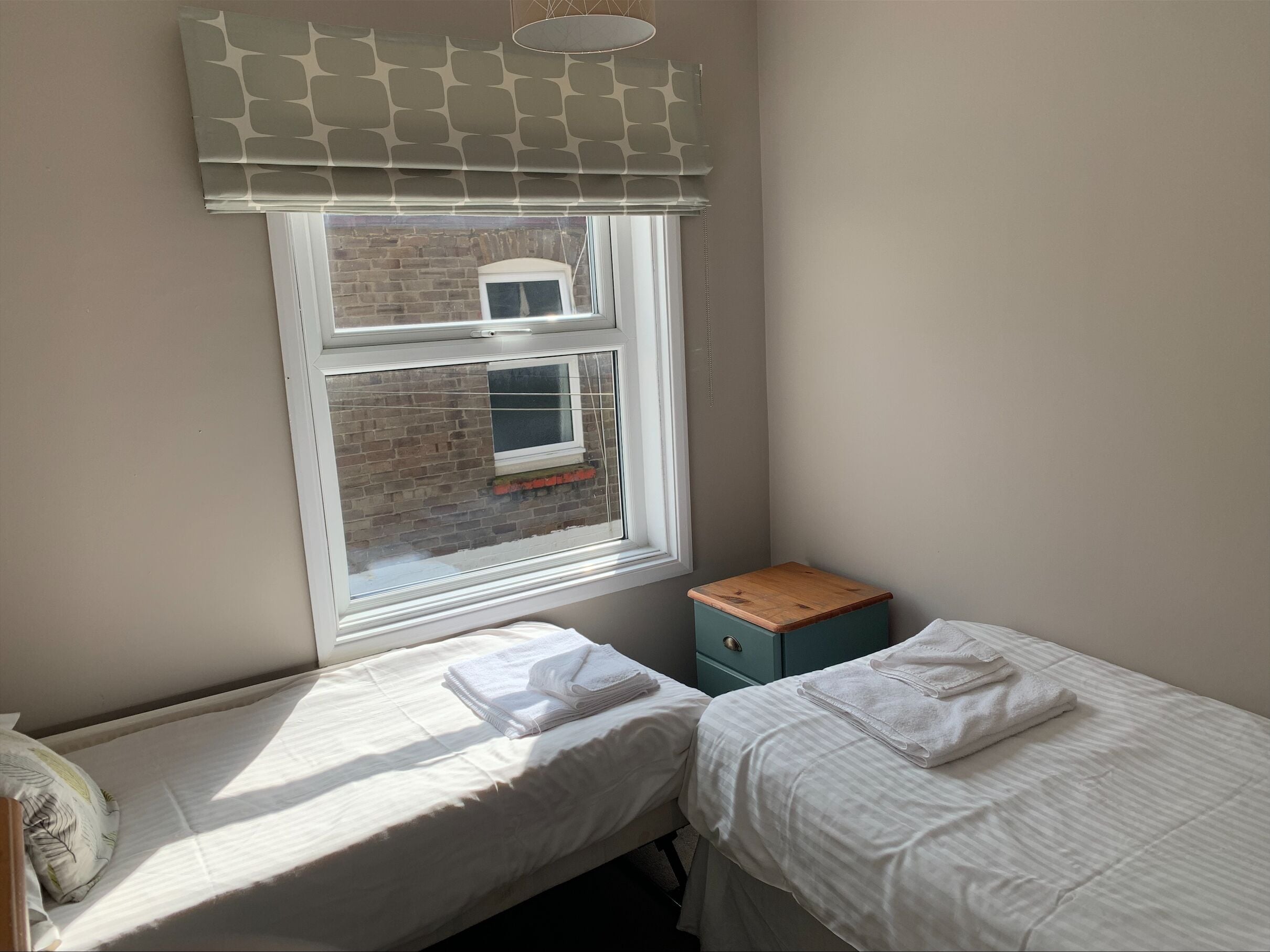 A small, tidy room with two single beds, a window with a view of a brick wall, and a small wooden cabinet.