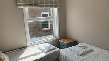 A small, tidy room with two single beds, a window with a view of a brick wall, and a small wooden cabinet.