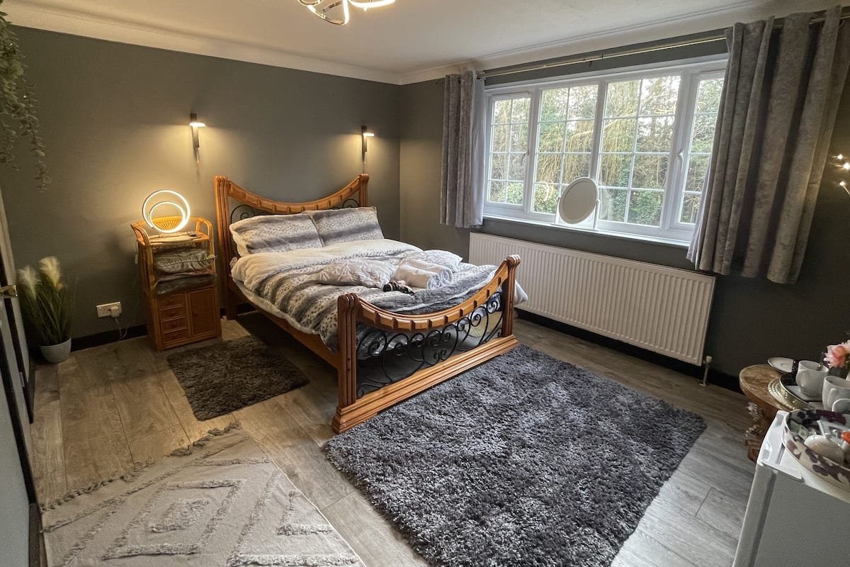 Un dormitorio con una cama de madera, lámparas de noche, un ventanal con cortinas, un radiador y una mesita con objetos decorativos.
