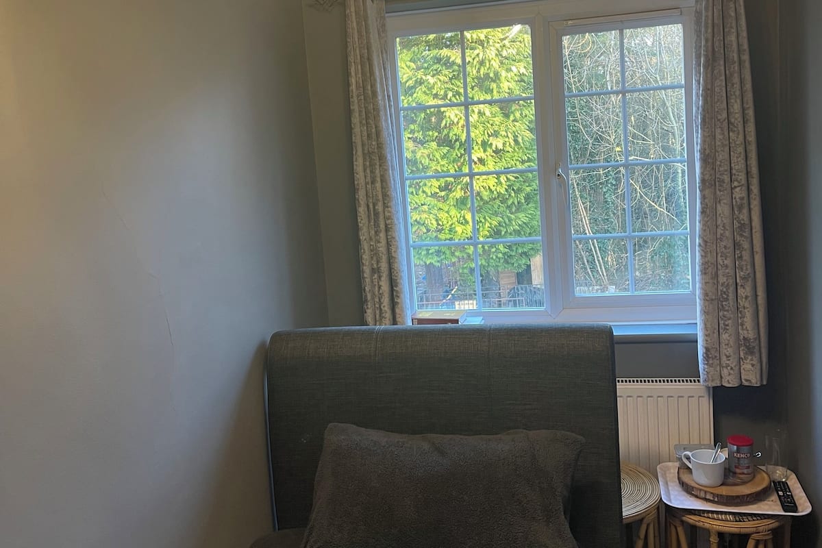 Un dormitorio con una cama, una silla, una mesita con una taza y un control remoto, y una ventana con cortinas.