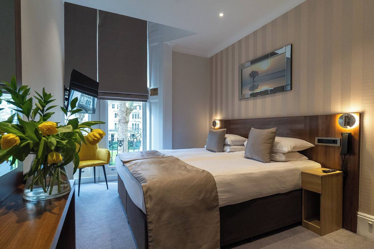 A modern hotel room with a large bed, bedside tables, a chair, a vase of flowers, and a city view.