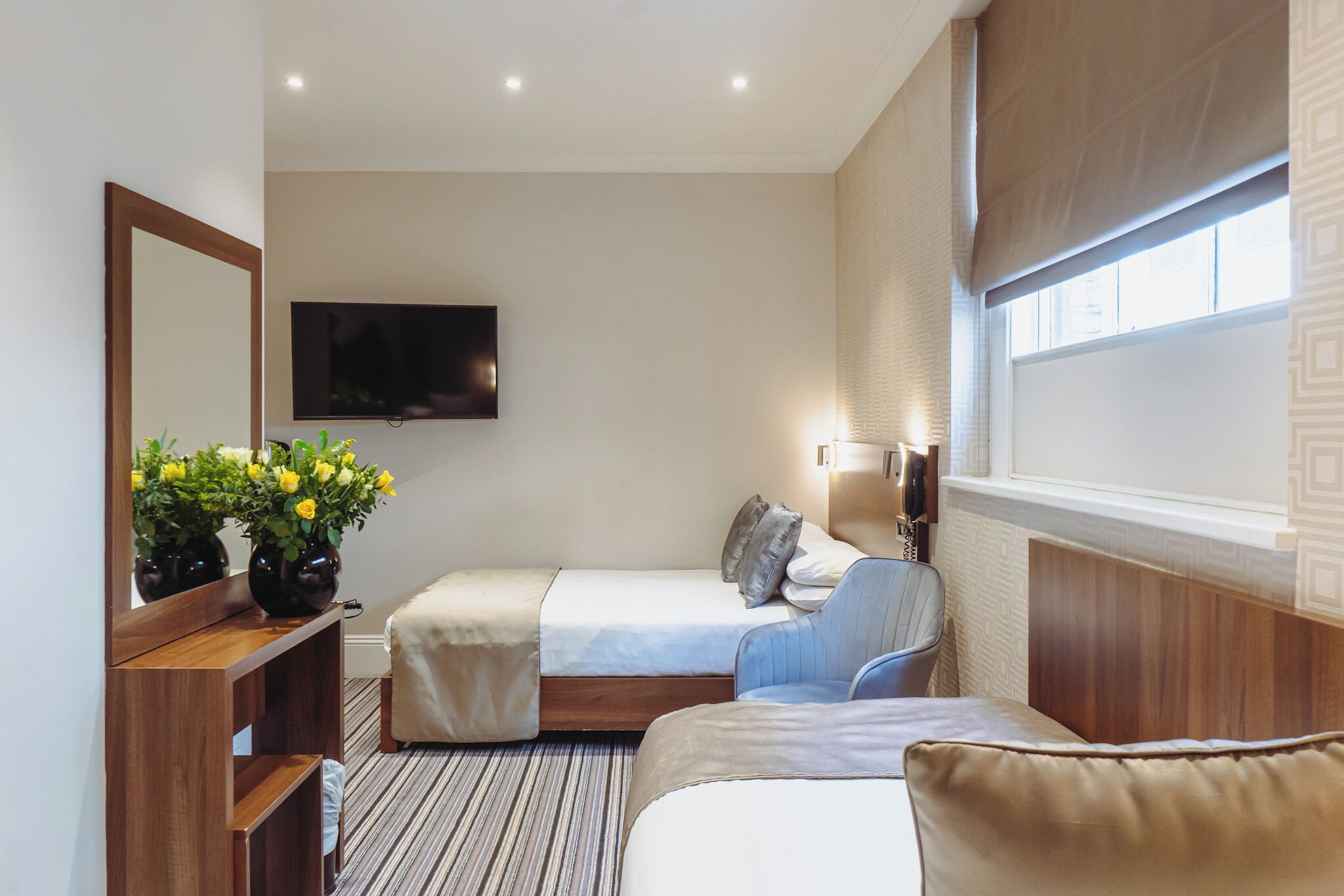 Habitación de hotel con dos camas, un televisor montado en la pared, una mesita de noche con un florero y una ventana con persianas.