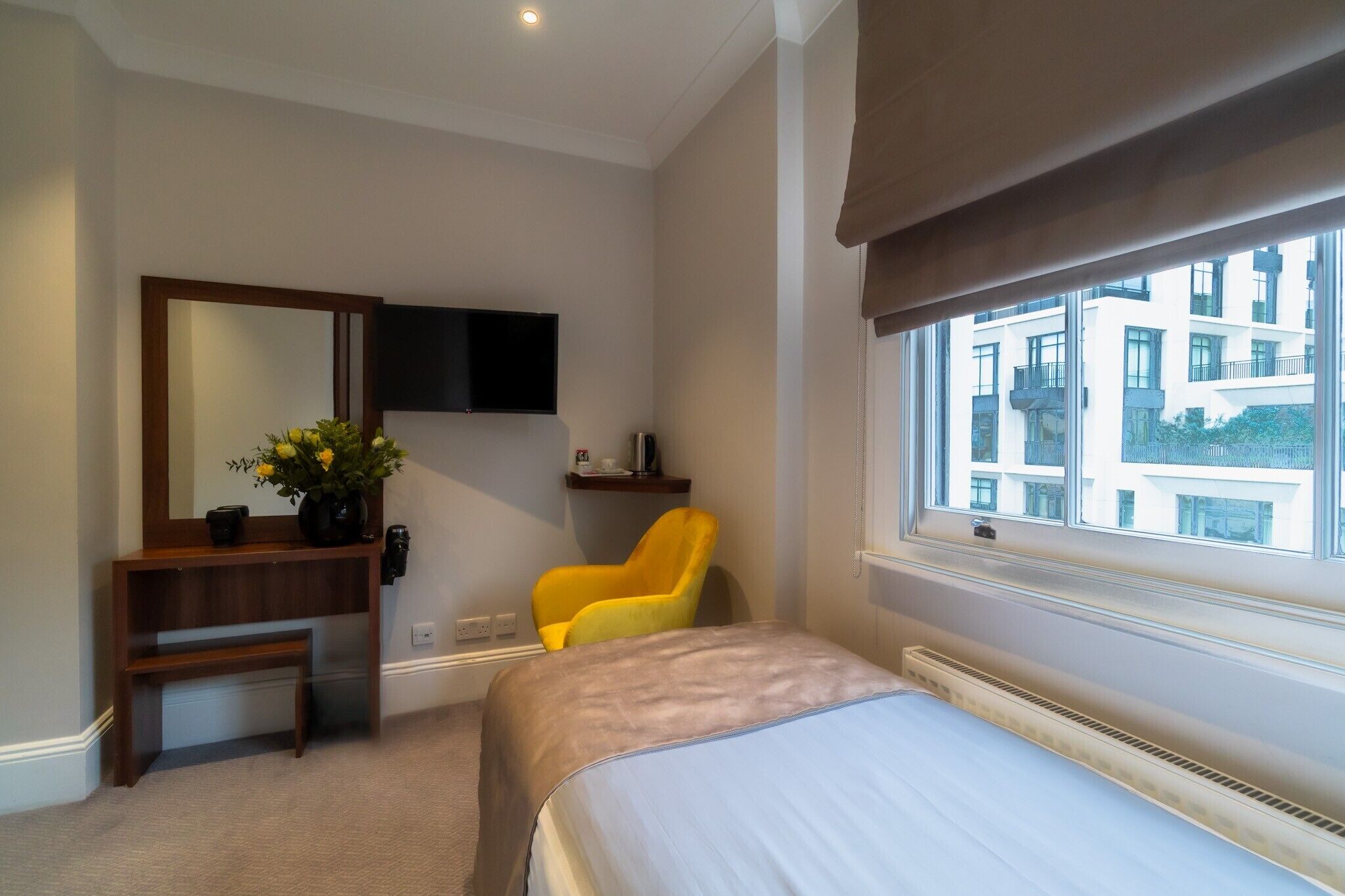 A hotel room with a bed, a desk with a mirror, a chair, a television, and a window with a view of buildings.