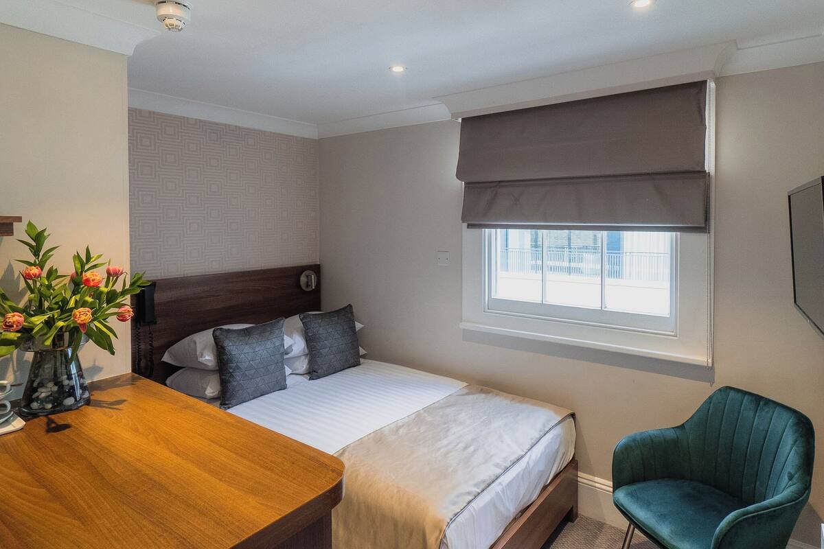 A hotel room with a bed, bedside table, chair, window with blinds, and a vase with flowers.