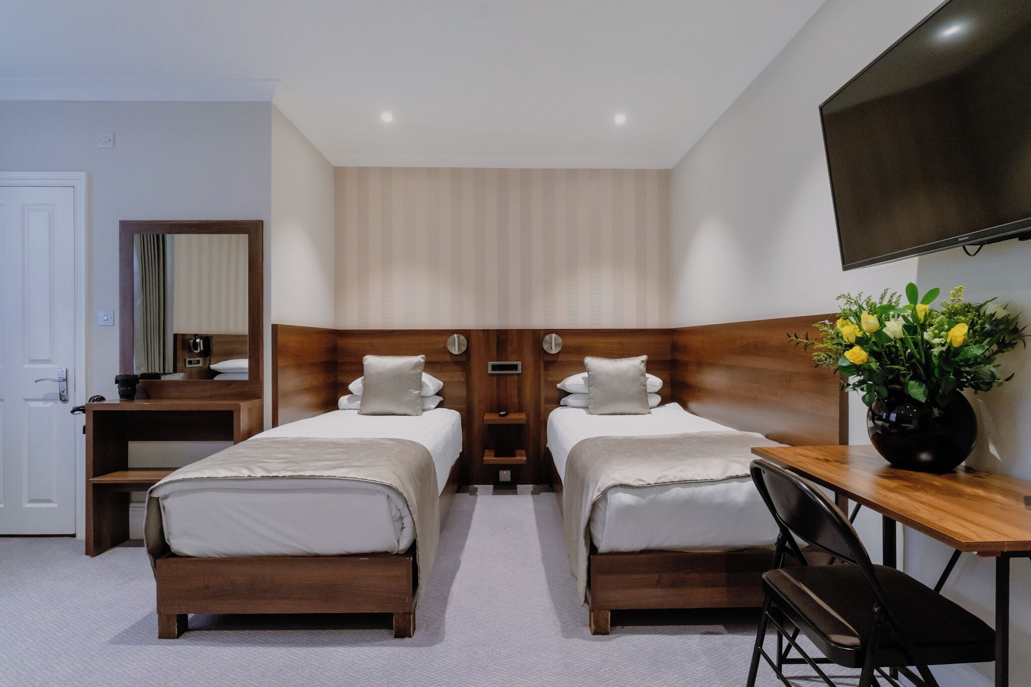 A hotel room with two beds, a desk, a chair, a TV, and a flower vase.