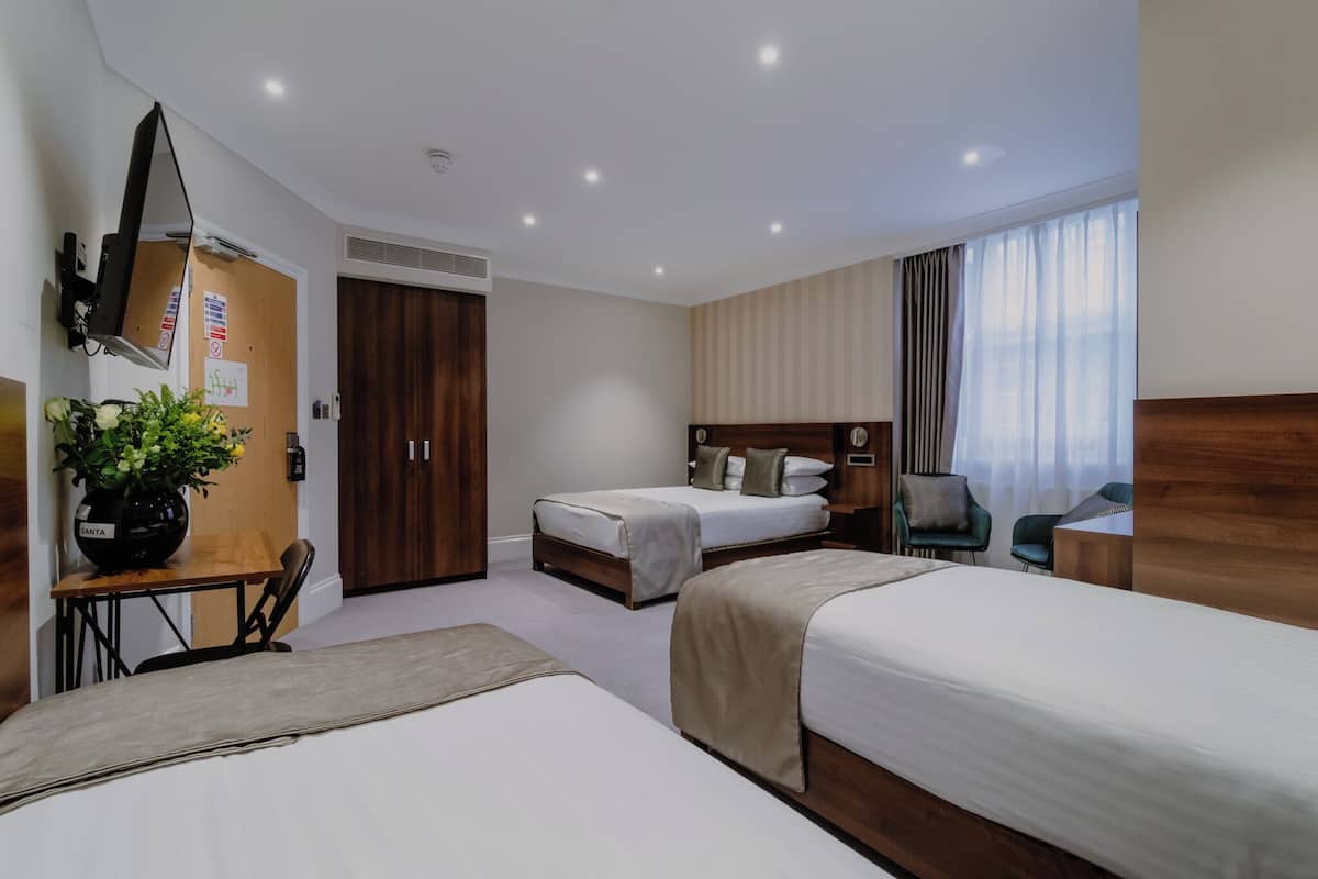 A hotel room with two beds, a desk with a chair, a potted plant, a TV, and a window with curtains.