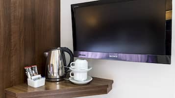 Un televisor de pantalla plana montado en la pared, una cafetera, una taza de café y un juego de cápsulas de café sobre un estante de madera.