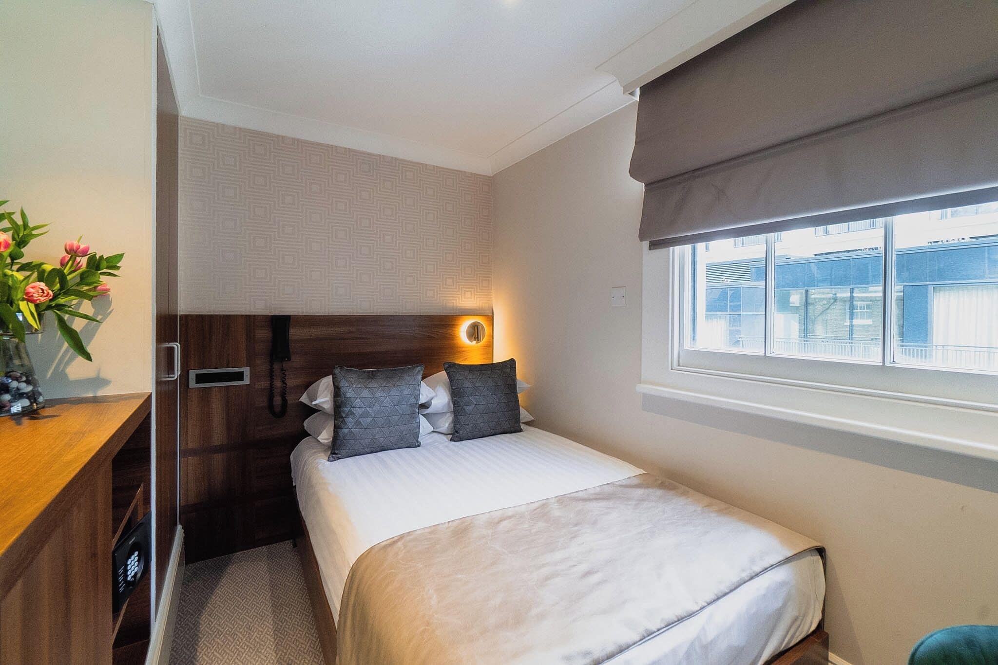 A compact hotel room with a bed, bedside table, and a window with a view of buildings.