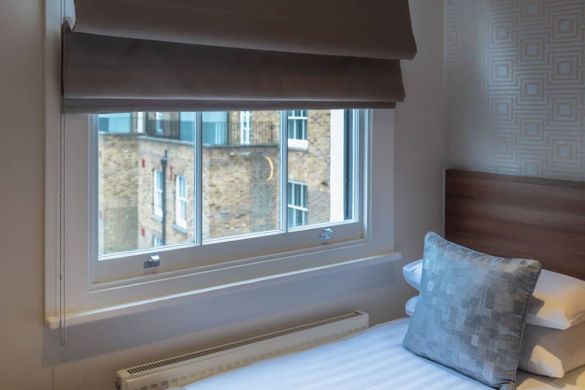 A bedroom with a large window, a bed with white linens, a grey pillow, and a brown ceiling blind.