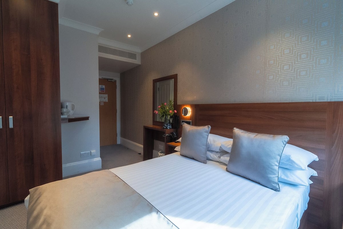 A hotel room with a large bed, wooden headboard, bedside table with a vase of flowers, and a mirror on the wall.