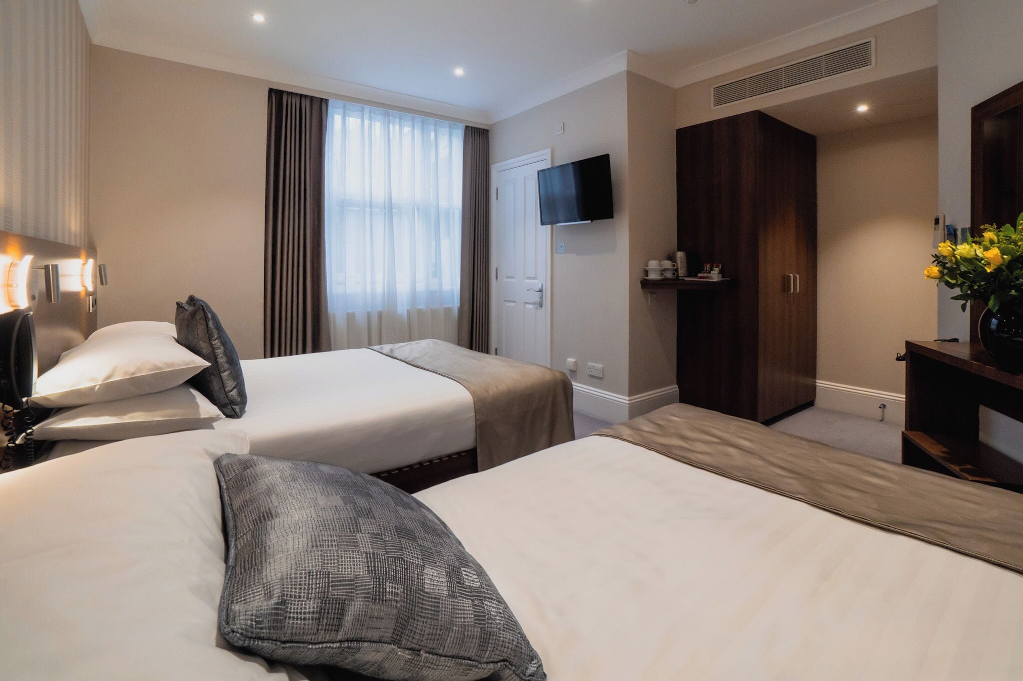A hotel room with two beds, a TV mounted on the wall, a wardrobe, and a desk with a vase of flowers.