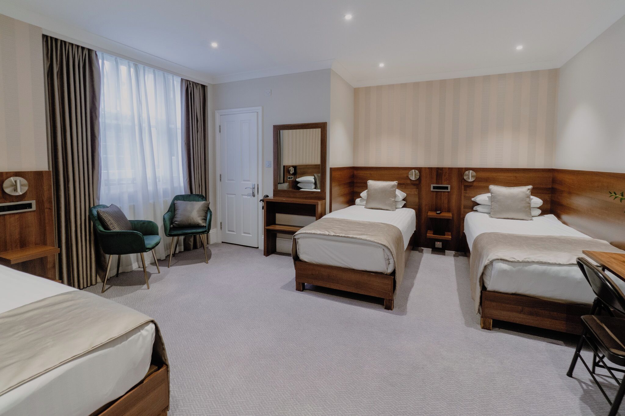 A hotel room with two single beds, a desk, a chair, a wardrobe, and a large window with curtains.