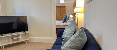 A living room with a TV on a stand, a blue sofa with cushions, a lamp, and a bed in the background.